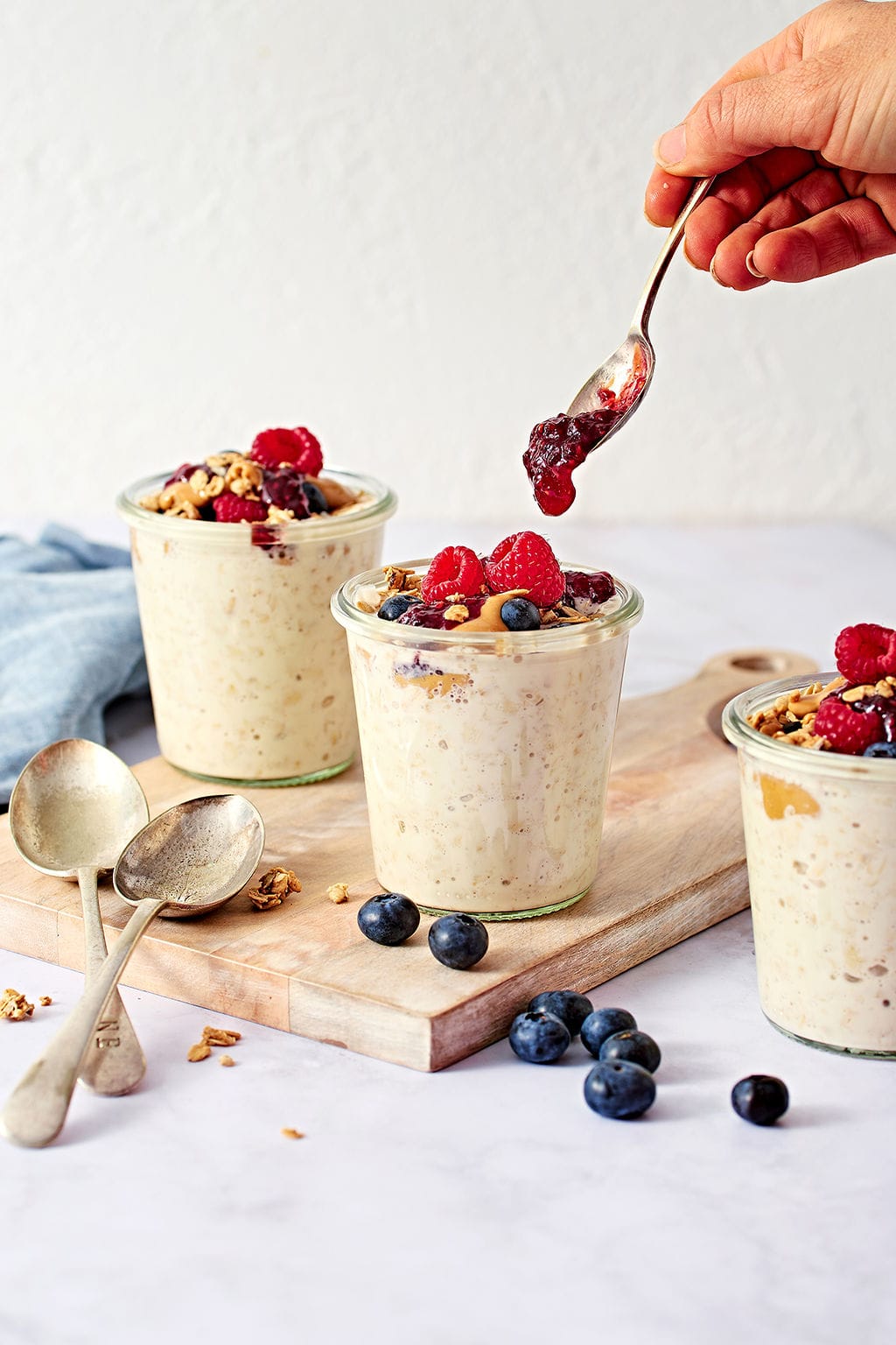 Creamy overnight oats in glass jars, being topped with fruit jam, perfect for breakfast or snacks.