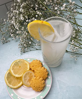 Lemonade with fresh lemon slices and biscuits, served with ice in a glass, on a light blue surface with white flowers in the background.