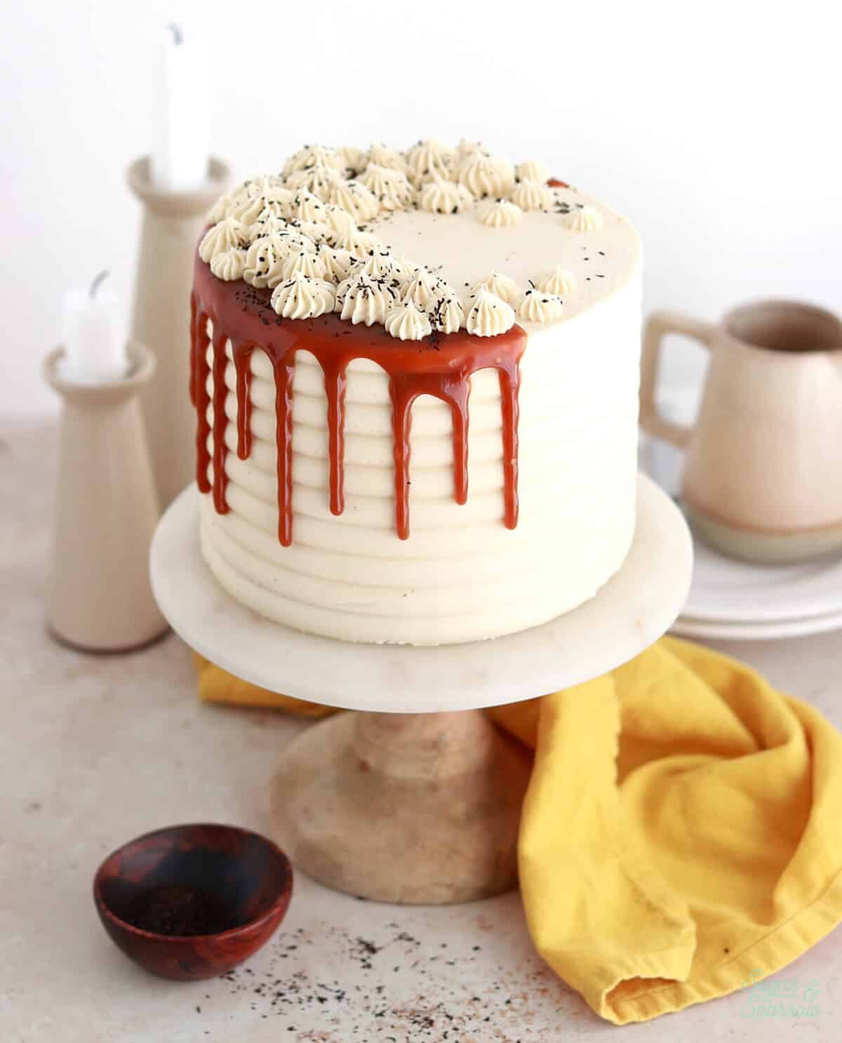 Elegant white cake with red drip glaze, topped with piped cream and sprinkles, on a rustic cake stand with a cosy kitchen background.