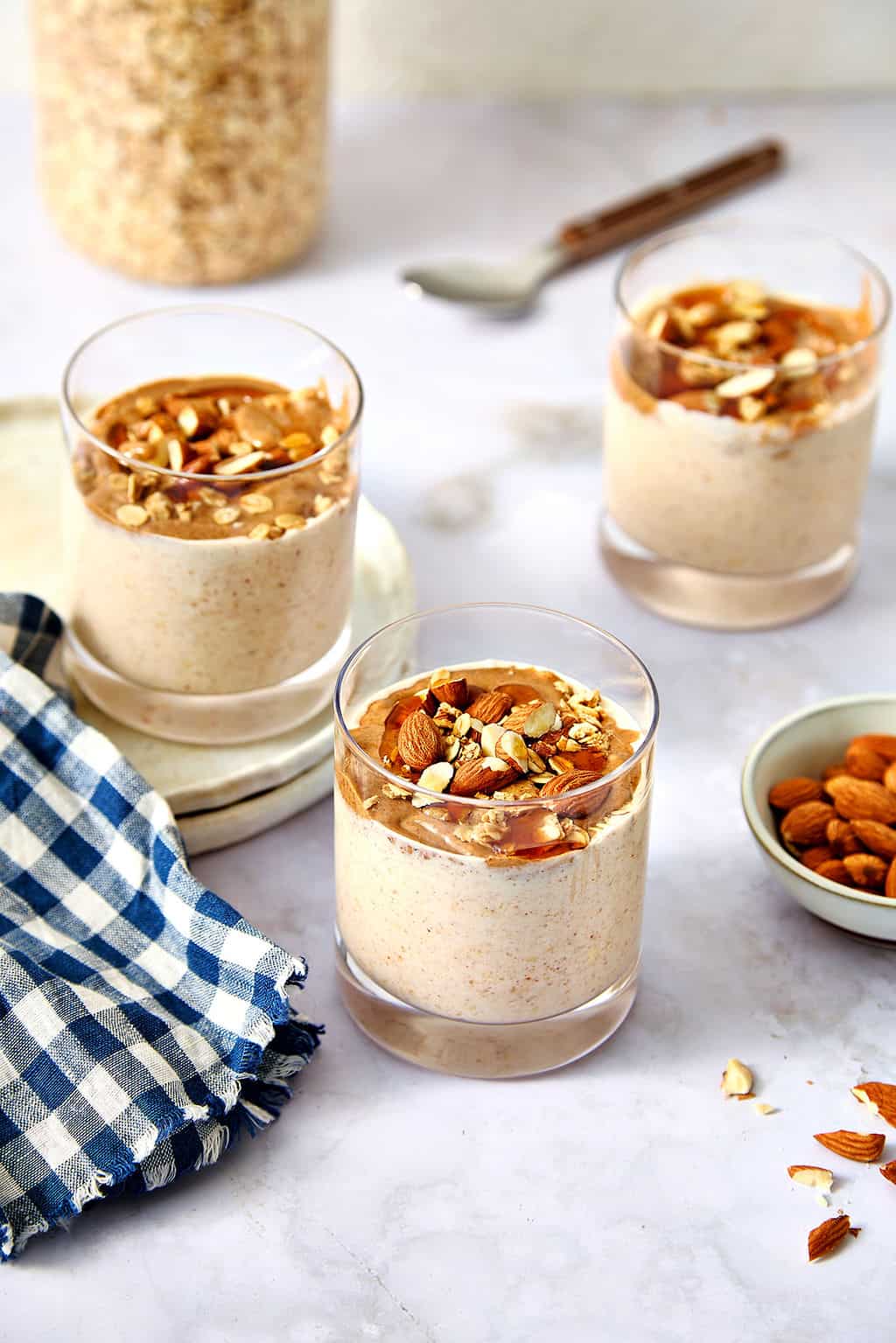 Creamy almond butter overnight oats topped with chopped nuts in clear glasses, sitting on a white marble bench top.
