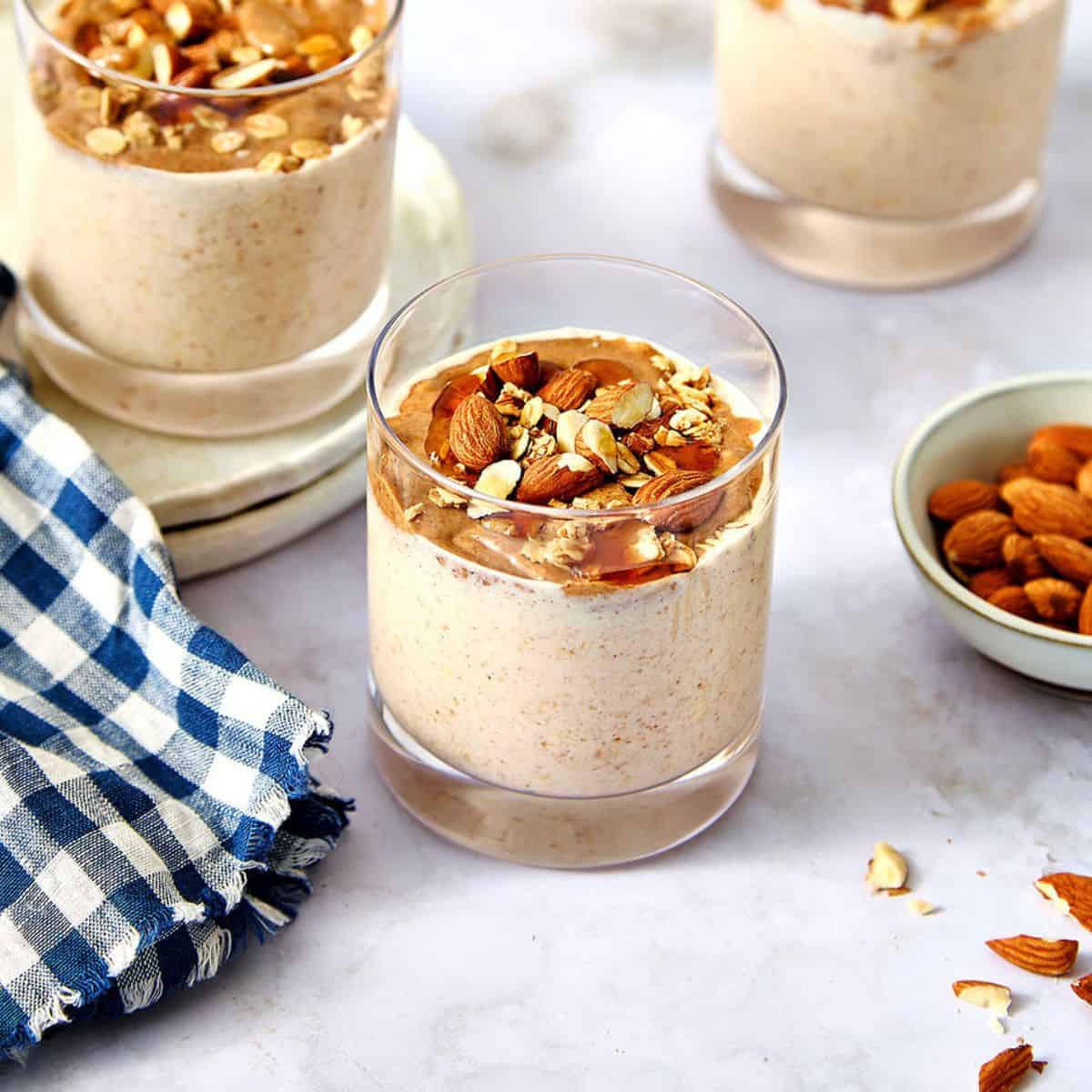 Creamy almond butter overnight oats topped with chopped nuts in a clear glass, served with a bowl of almonds and a blue checkered cloth on a marble table.