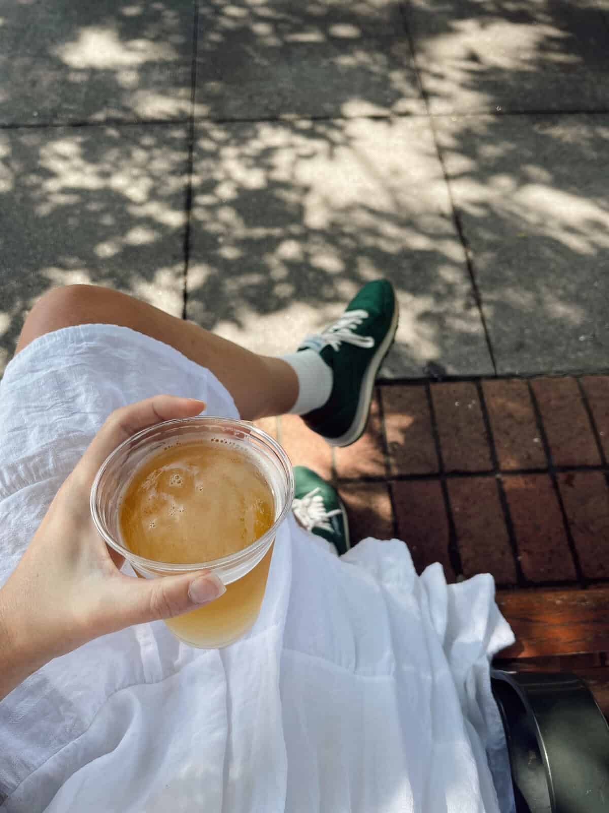 Person enjoying a cold beverage outdoors on a sunny day, sitting on a bench with a view of shaded pavement and brick walkway.