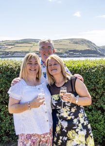Three people smiling outdoors holding drinks with countryside and coastal hills in the background