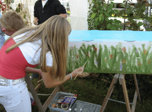 Children painting a nature scene on a large canvas at an outdoor art workshop.