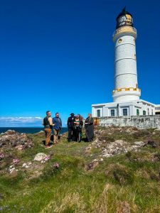 Group of people near a lighthouse on a grassy landscape with rocks and flowers.