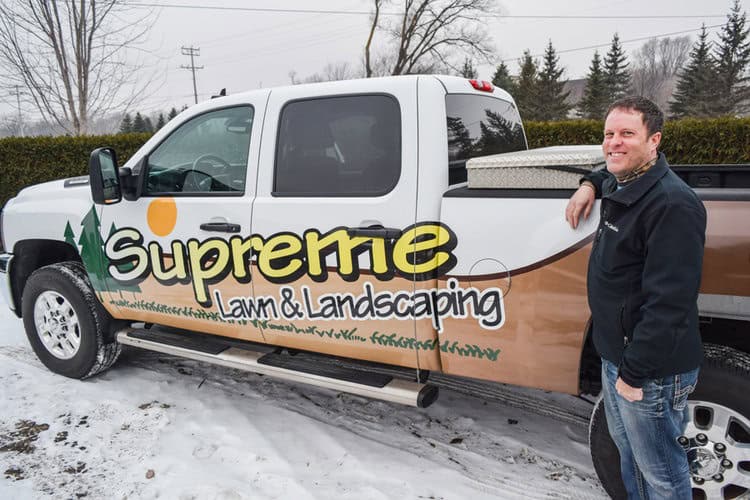 Supreme lawn landscaping truck with company logo and smiling man in winter setting.