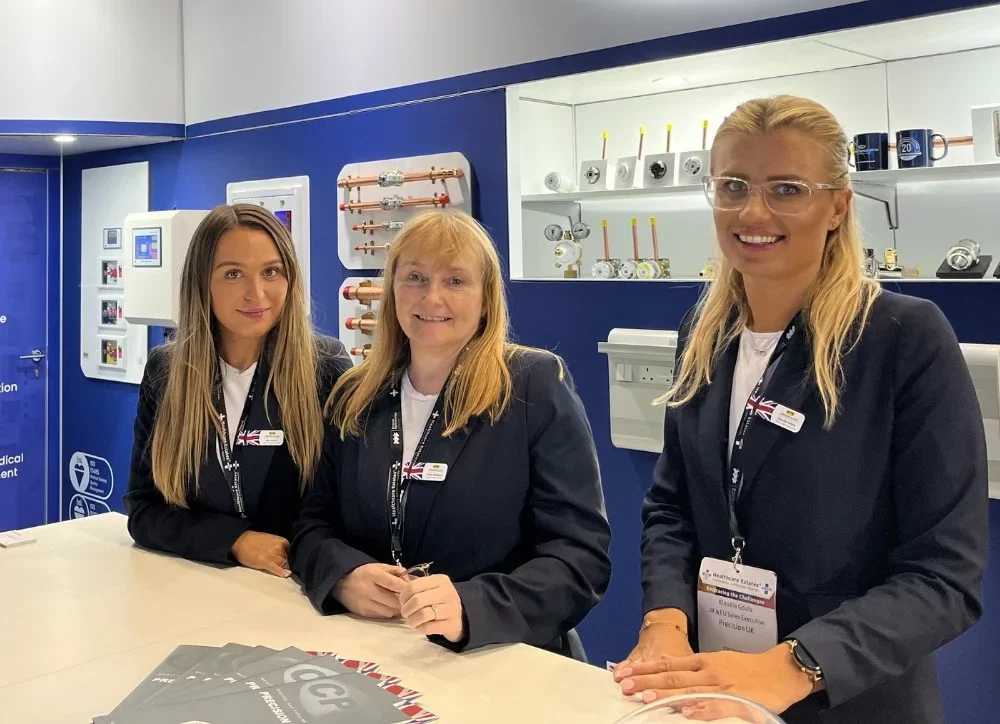 Precision UK team on the desk at Healthcare Estates exhibition. Navy CPX logo in the background with various medical gas pipeline equipment lit up on display shelves