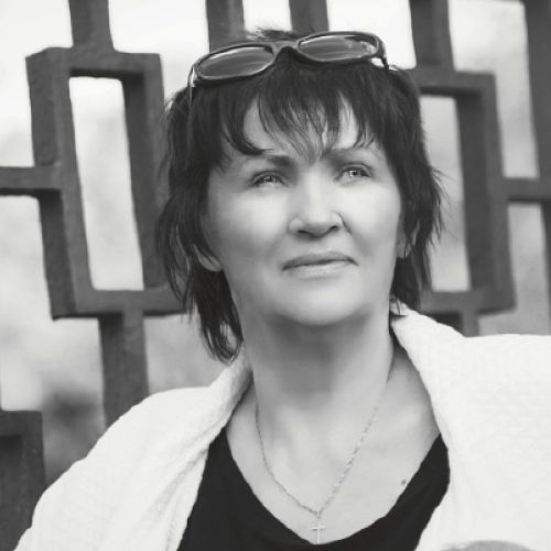 Pokojná žena s brýlemi na hlavě, fotografovaná v černobílém provedení, v pozadí umělecká železná konstrukce, symbol zodpovědnosti za vodu a životní prostředí.
