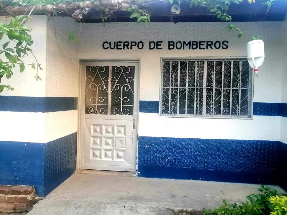 Puerta de entrada del cuerpo de bomberos en Huila, con rejas metálicas y señalización visible, en un edificio con paredes pintadas en azul y blanco en Huila TV.