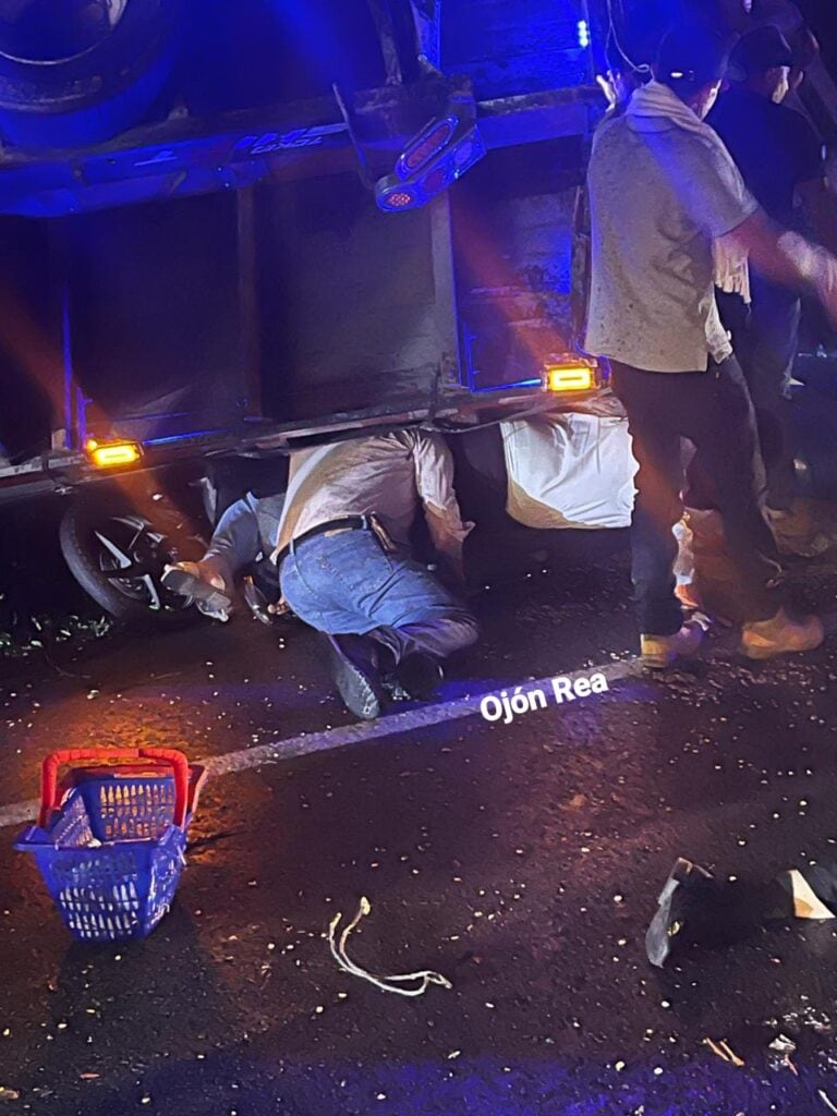 Accidente de tránsito en Ojón Rea con personas revisando debajo de un vehículo, iluminación nocturna, presencia de una cesta de supermercado y cubrebocas, escena de emergencia o accidente en la vía.