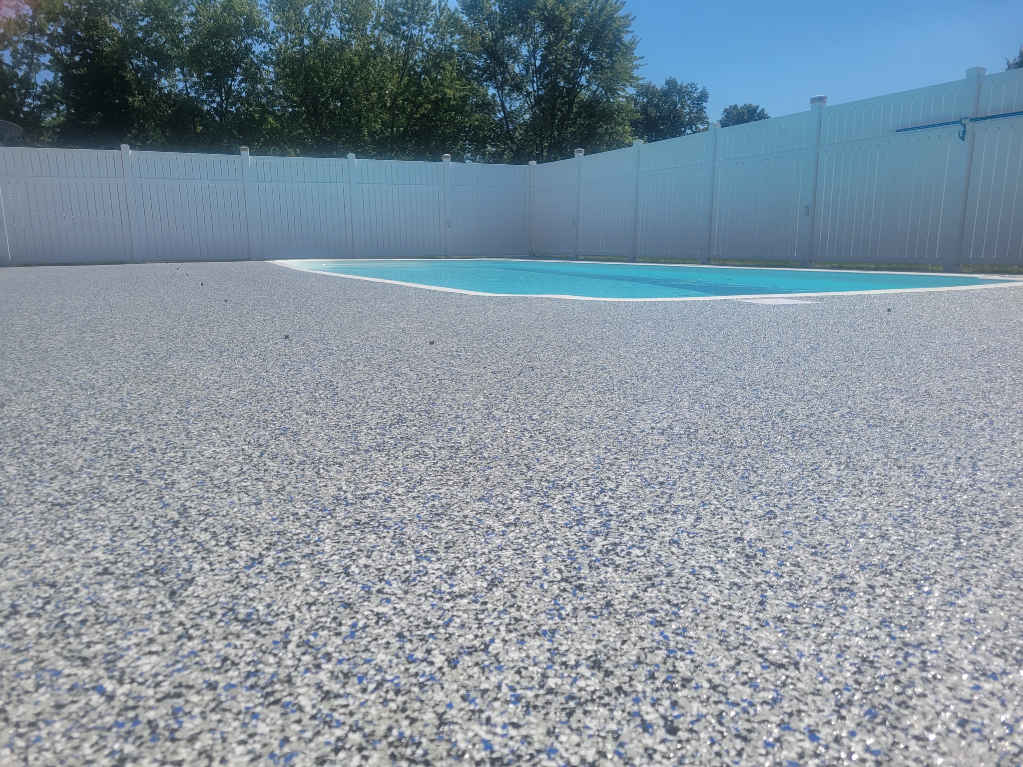 The image shows a backyard pool area finished by J&P Coatings. The concrete deck looks safe and inviting for families to enjoy spending time outdoors. The yard is private with a tall fence and has green trees around it.