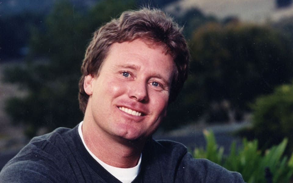 Portrait of a smiling man with blond hair, wearing a dark sweater, holding a book titled "Don't Sweat The Small Stuff & It's All Small Stuff," with a natural, hilly landscape visible