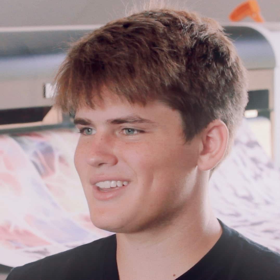 A young man with tousled brown hair and a slight smile looks to his right in a room with blurred artistic wall décor in the background, embodying the essence of Generation Freedom.