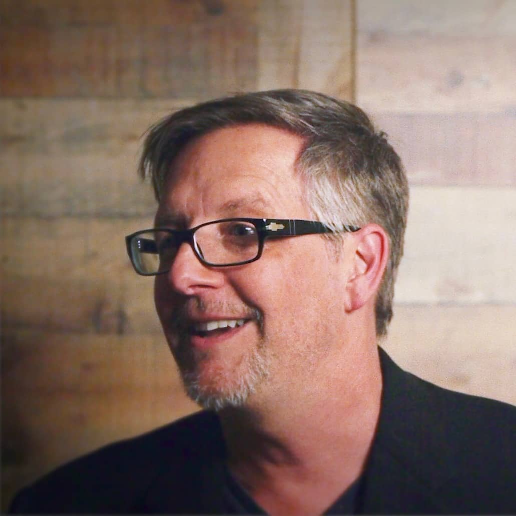 A middle-aged man with glasses smiling slightly, looking off to the side against a blurred wooden background. As part of Generation Freedom, he has short, graying hair and is wearing a black shirt.
