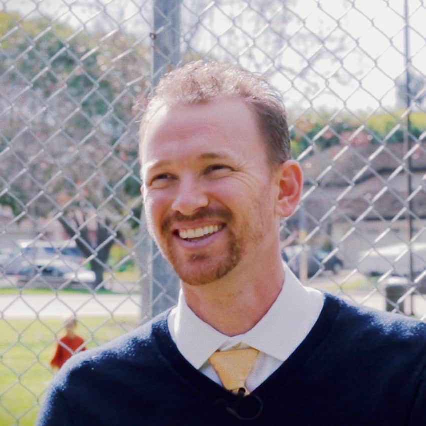 A smiling man from Generation Freedom with short blond hair, wearing a navy sweater over a collared shirt and tie, stands in front of a chain-link fence in a sunny outdoor setting.