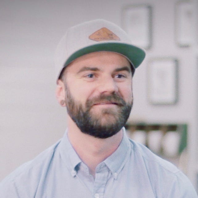 A smiling man with a beard, wearing a gray cap and light blue shirt, looks slightly to the side. He's in a softly blurred indoor setting adorned with framed pictures that celebrate Generation Freedom in the