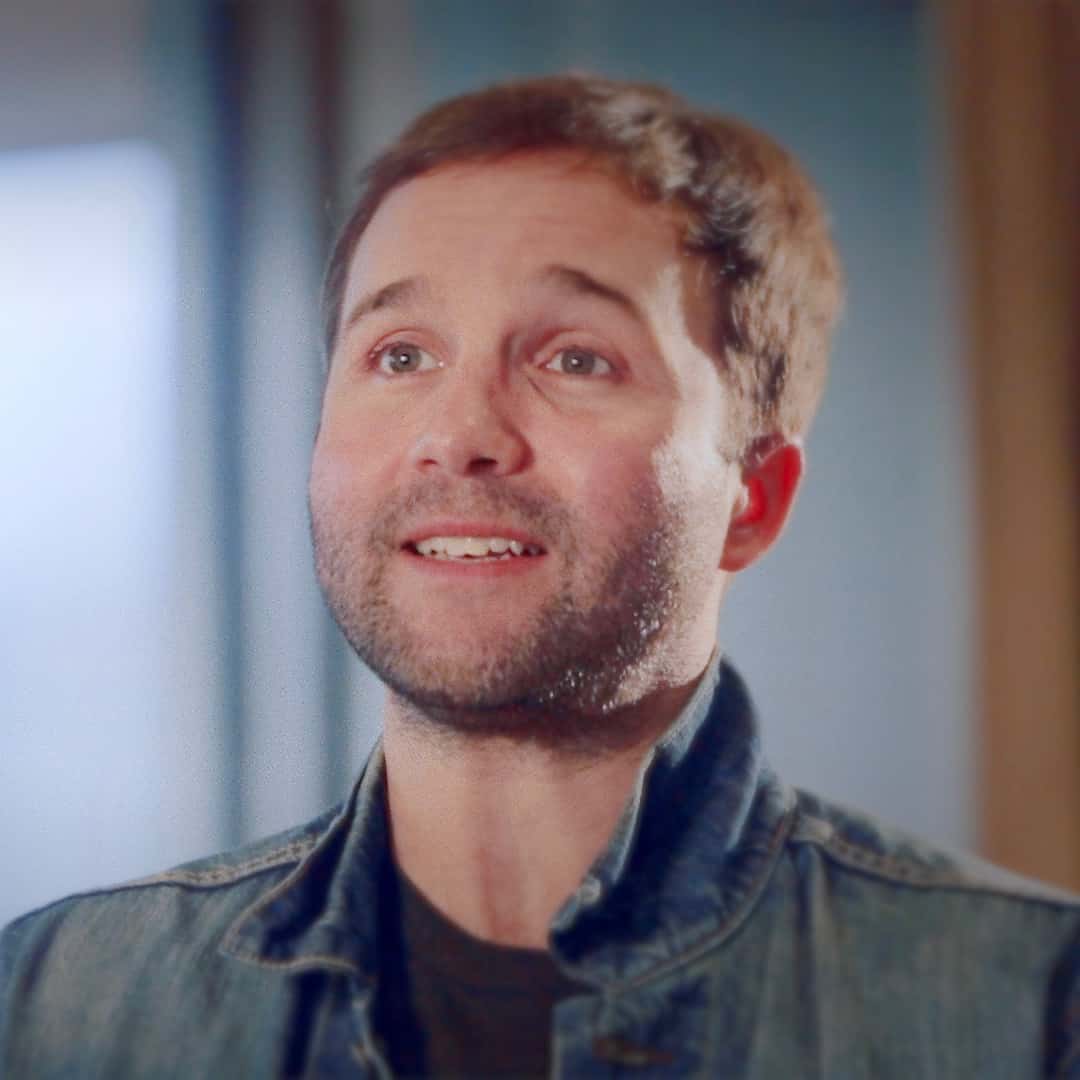 A man with a beard smiling slightly, looking upwards with an expression of hope or anticipation. He is wearing a denim jacket, set against a softly blurred background, embodying the spirit of Generation Freedom.