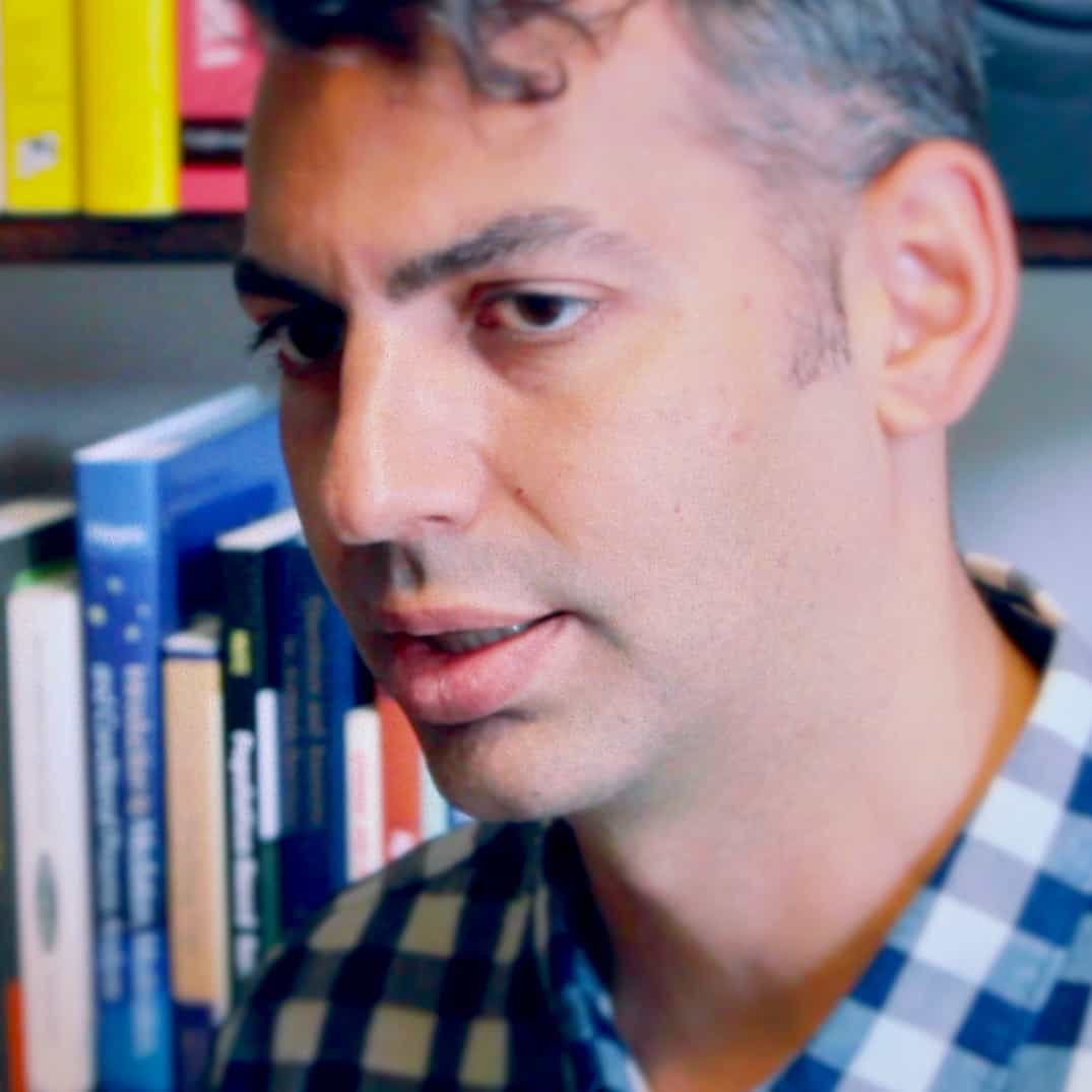 Close-up of a man with dark hair and a thoughtful expression, wearing a plaid shirt, standing in front of a bookshelf filled with books from Generation Freedom.