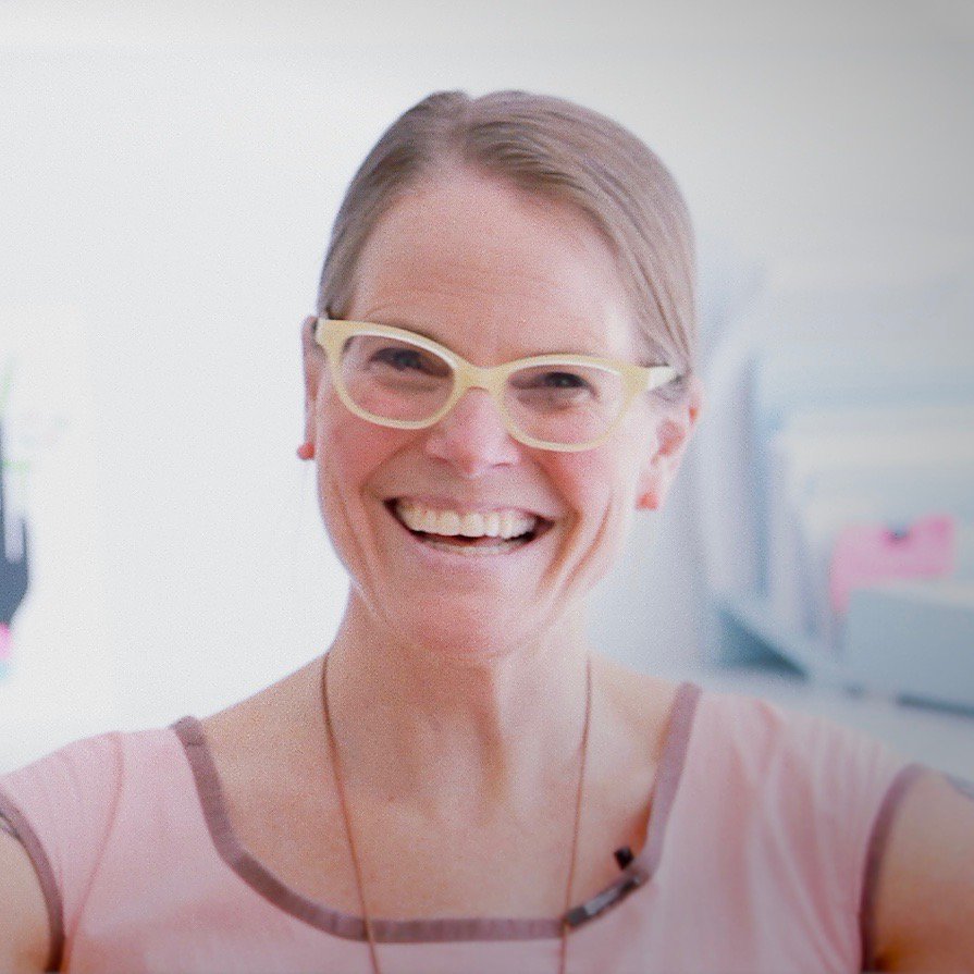 A woman with a broad smile, wearing yellow-framed glasses and a pink top, stands in a brightly lit room, embodying the spirit of Generation Freedom.