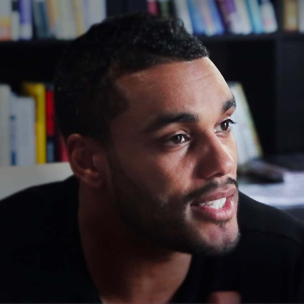 Close-up of a young man from Generation Freedom with short curly hair and light stubble, smiling gently during a conversational interview, with a blurred bookshelf in the background.