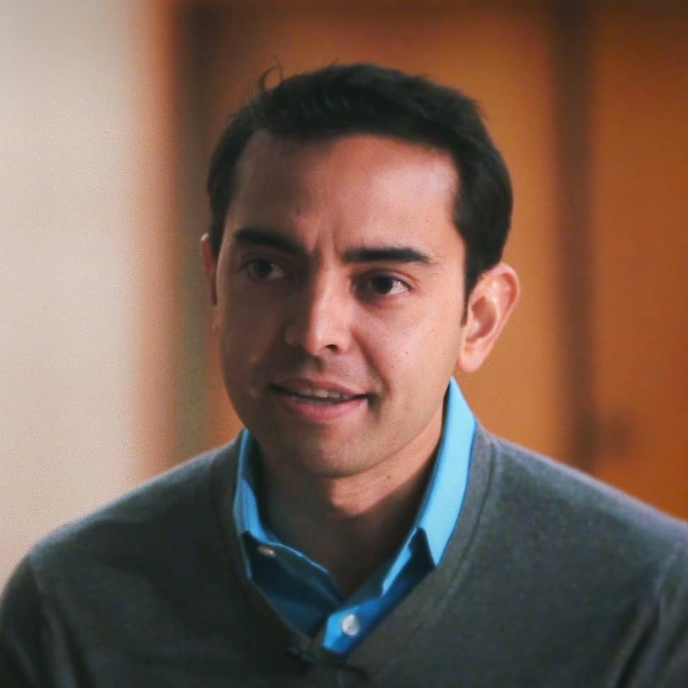 A portrait of a man with dark hair and prominent eyebrows, wearing a gray sweater over a light blue shirt. He has a focused expression with a slight hint of concern, embodying the resilience of Generation