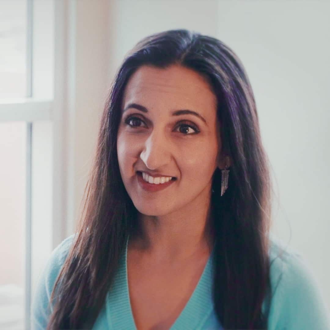 A woman with long dark hair, wearing a turquoise top and dangling earrings, smiles gently in a warmly lit indoor setting, epitomizing Generation Freedom.