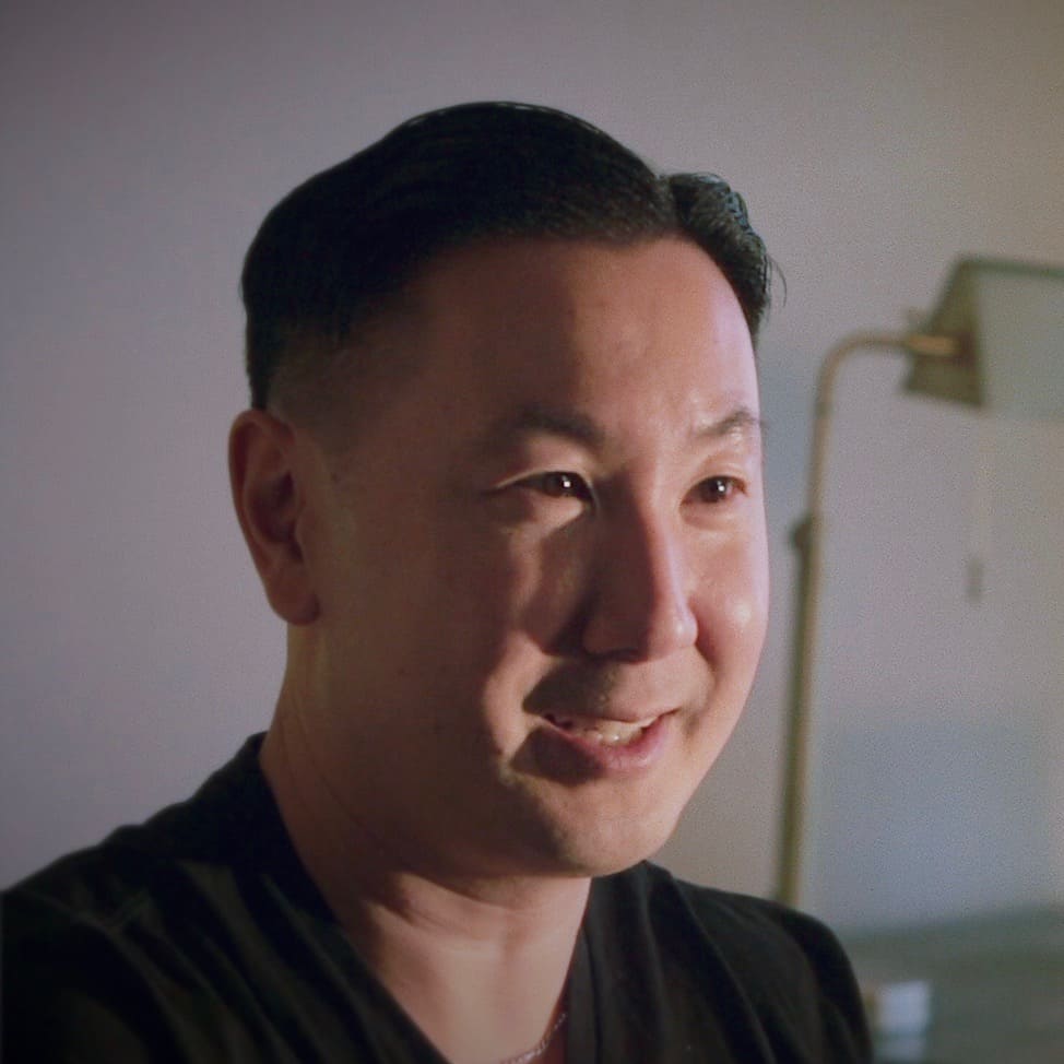 Close-up of a smiling man with short dark hair styled in a wave, wearing a black "Generation Freedom" t-shirt, against a softly lit indoor background.