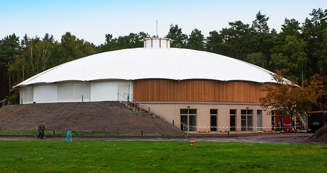 Moderne Veranstaltungshalle mit weißem, gewölbtem Dach und Holz-Fassadenelementen in einem grünen Park, perfekt für Events, Konferenzen und kulturelle Veranstaltungen in Deutschland.