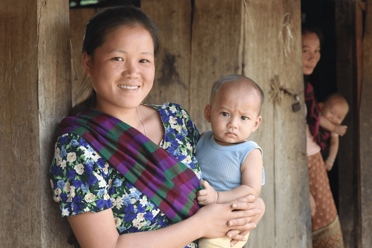 Fröhliche asiatische Mutter mit Kleinkind vor traditionellem Holzhaus in ländlichem Gebiet, Nahaufnahme, Nahaufnahme, Menschen, Familienleben, Gemeinschaft, Entwicklung, Erfolg, Liebe, Glück, Familienbindung, nachhaltige Entwicklung, soziales Engagement, ländliche Gemeinschaft, Kultur, Indonesien.
