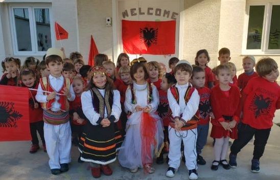 Alttag: Gruppe von Kindern in traditionellen albanischen Trachten vor einem Schulgebäude mit albanischer Flagge, bei einer kulturellen Veranstaltung.