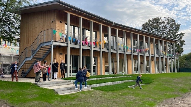 Moderne Holzkonstruktion mit großen Fenstern und bunten Fahnen, umgeben von grüner Rasenfläche, mit Menschen, die das Gebäude erkunden und Kinder, die draußen spielen, bei Tageslicht.