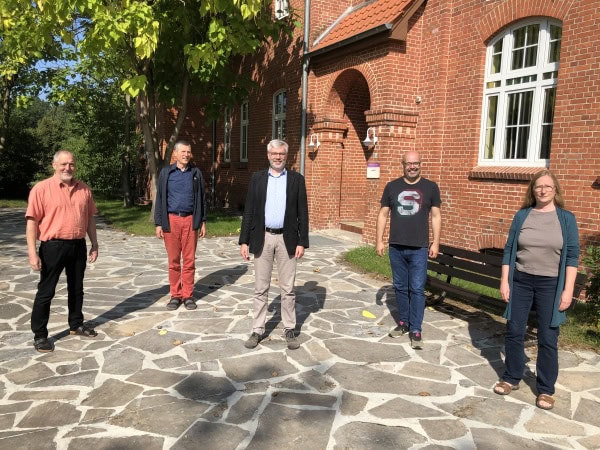 1. Vielfältiges Team vor rotziegeligem Gebäude bei Sonnenlicht, Außenaufnahme, professionelle Atmosphäre, Kreativität, Zusammenarbeit, Innovation, freundliche Stimmung, admintiv, modern.