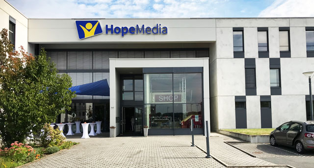 Hoffnungsmedien Firmengebäude mit HopeMedia Logo, moderne Architektur, Empfangsbereich, Büroumgebung, Symbol für Medienarbeit, Standort in Deutschland.