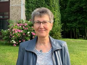 Erwachsene Frau im Freien, lächelnd, mit Brille, vor grüner Natur und Blumen, repräsentiert Bildung und Umwelt, passend für religiöse oder gesellige Veranstaltungen.