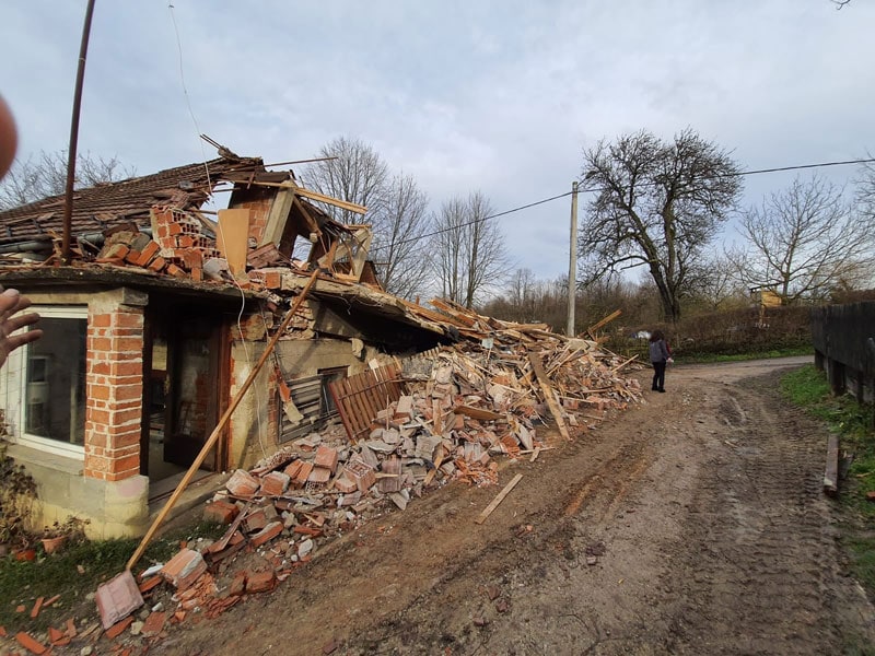 Zerstörtes Haus nach Unfall, beschädigtes Gebäude, Trümmer und Bruch, mit zwei Personen im Hintergrund, unwegsame Straße, Natur im ländlichen Gebiet, Schadensbild nach Katastrophe, Notfall, Rettungseinsatz, Sicherheitsrisiko, Notstand, Schadensanalyse.