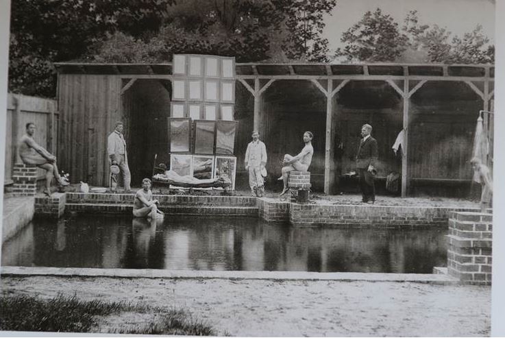 Blick auf eine historische Szene mit mehreren Personen am Wasser in einem Holzschuppen, im Hintergrund Bäume, vermutlich ein altes Theater- oder Freizeitfoto, passend für historische und kulturelle Themen.