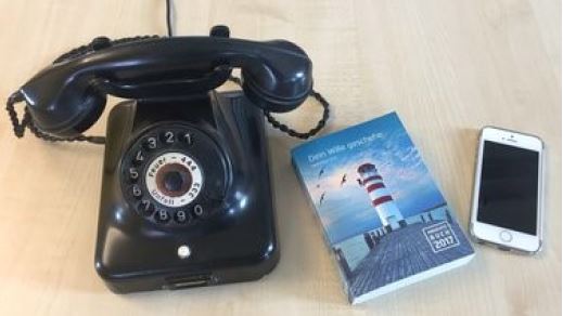 Alte schwarze Wählscheiben-Telefon mit Hörer neben einem Buch und Smartphone auf Holztisch.