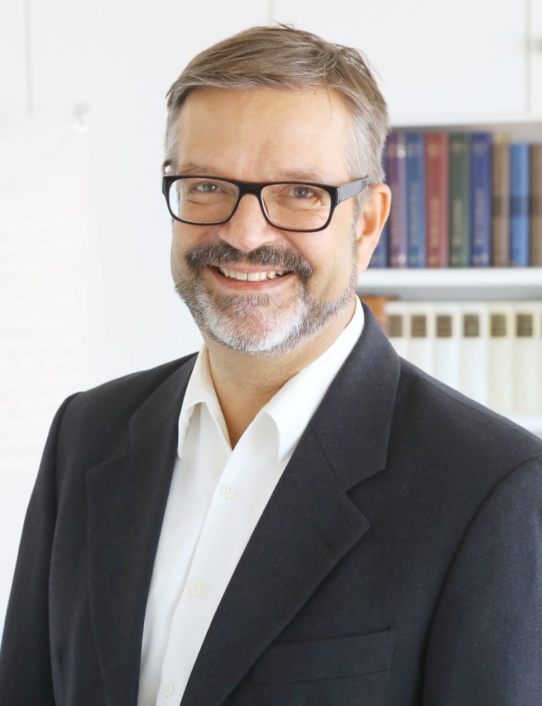 Freundlicher Mann im Geschäftsanzug mit Brille vor Bücherregal, symbolisiert Kompetenz und Vertrauen in der Verlagsbranche Advent-Verlag, ideal für christliche Literatur, Kirchenbedarf & Adventszeit-Produkte, professionelles Portrait.