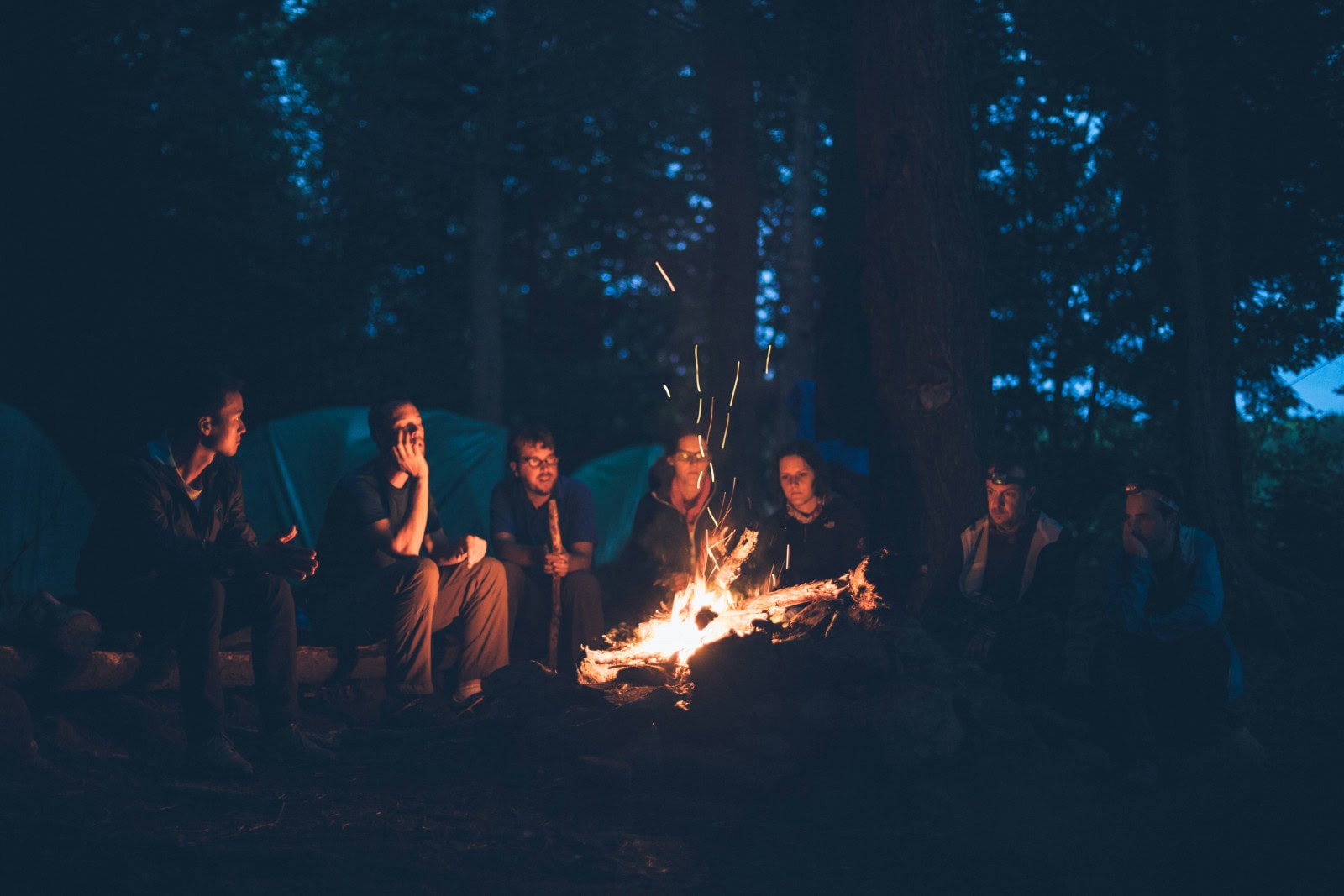 Lagerfeuer mit Gruppe von Menschen in einem Wald bei Nacht, entspannte Atmosphäre, Gemeinschaft, Outdoor-Aktivitäten, Natur, Freunde, Camping, Abendstimmung, dunkler Himmel.