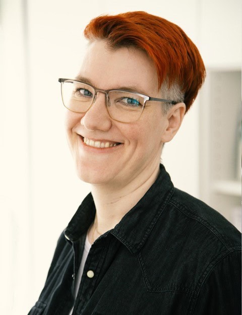 Brennende rote Haare, Brille, freundliches Lächeln, Porträt einer Frau im Büro, moderne Arbeitsumgebung, authentisch, professionell, Fokus auf Persönlichkeit, natürliches Licht.