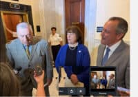 Older man in a gray plaid suit being interviewed by reporters in a marble-floored lobby, with a woman in blue blazer and a security guard in the background.