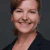 Smiling woman in a dark blazer and blue top, posing for a professional headshot against a neutral gray background.