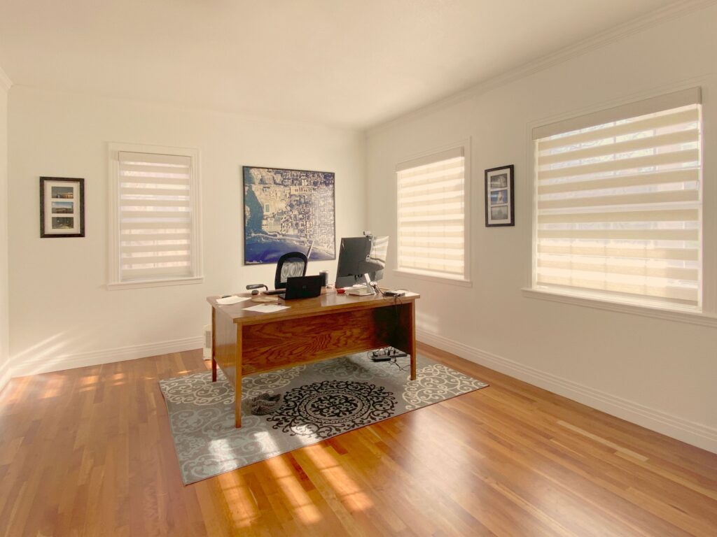 Window shades in a bright home office with wooden desk and artwork.