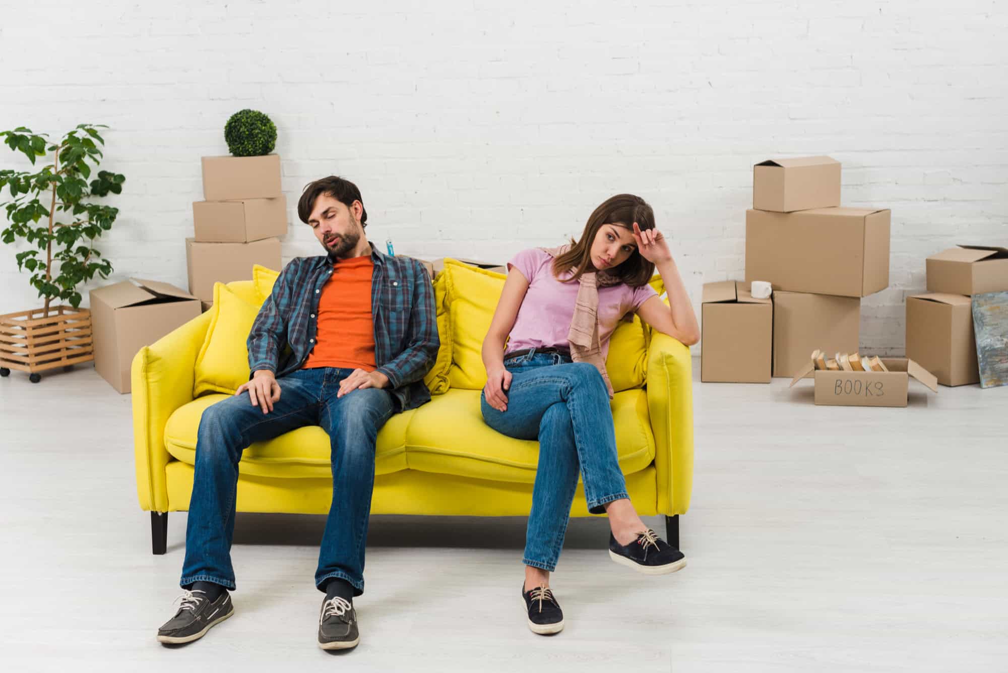Frustrated couple sitting on yellow sofa amidst moving boxes.
