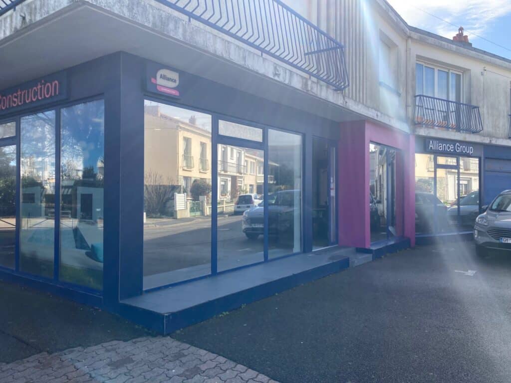 agence-lessables Façade d'une agence Alliance Group avec vitrines modernes et un habillage en rose et bleu, située dans un environnement urbain avec des places de stationnement à proximité.