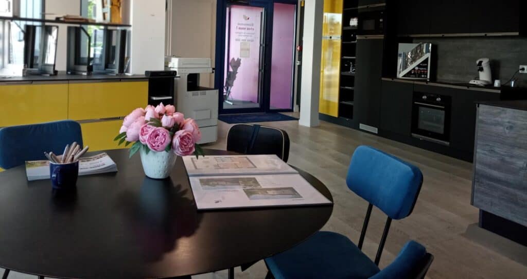 agence-lessables Intérieur d’une agence Alliance Group avec un espace de réunion chaleureux, une table ronde décorée d’un vase de fleurs roses, des plans de projets posés sur la table, et une cuisine intégrée moderne en arrière-plan avec des façades noires et jaunes.