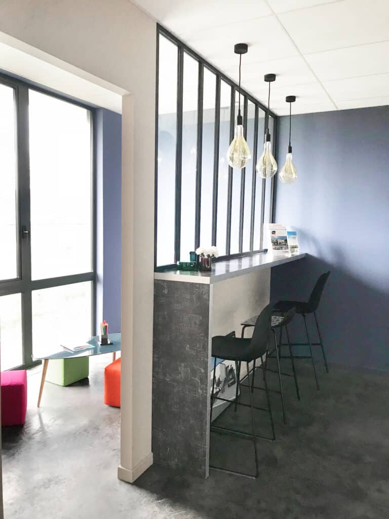 Espace intérieur moderne et lumineux avec verrière industrielle, murs bleu foncé, mobilier design incluant des tabourets hauts et une table basse colorée, créant une ambiance chaleureuse et conviviale.