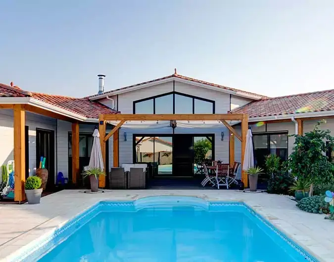 Maison moderne avec toit en tuiles, large terrasse en bois, et piscine entourée de verdure, sous un ciel dégagé et ensoleillé.