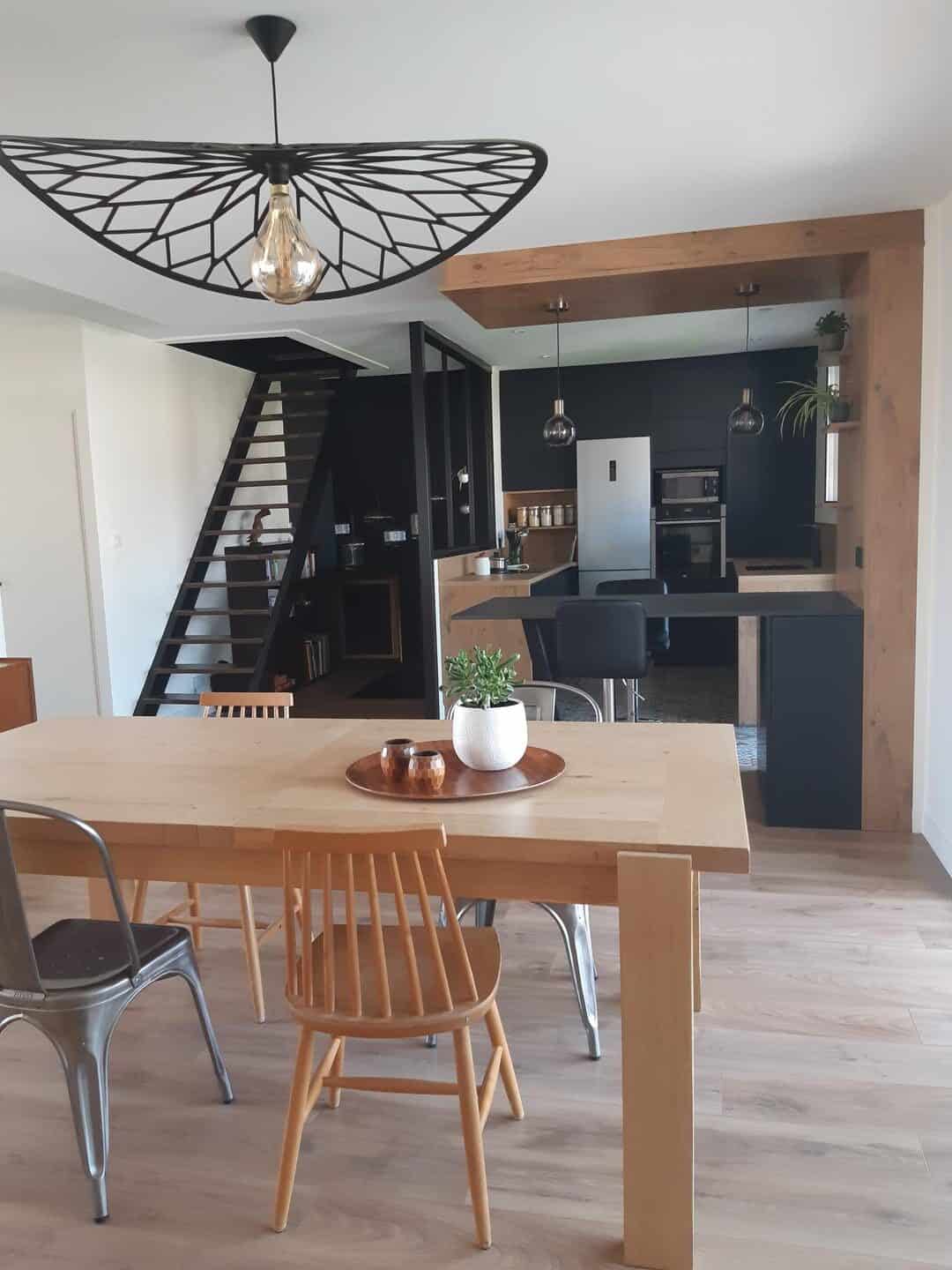 Salle à manger ouverte sur une cuisine moderne, avec une table en bois clair, des chaises design en bois et métal, et un luminaire suspendu au style contemporain. L’ensemble crée une ambiance chaleureuse et conviviale.