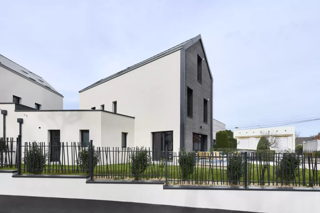 Maison moderne à trois niveaux avec façade en enduit clair et bardage sombre, entourée d’un jardin clôturé, sous un ciel nuageux. HD